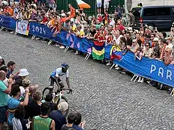 Ein Rennradfahrer fährt eine bergab führende, mit Kopfstein gepflasterte Straße hinunter an der beidseitig hinter Banden Publikum zusieht.