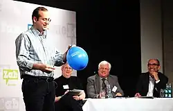 Ein Mann steht mit einem blauen Luftballon auf einer Bühne. Im Hintergrund sitzt eine Jury aus zwei Männern und einer Frau an einem Tisch