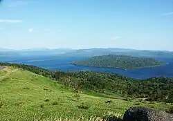 Blick von einem Gras bewachsenen Hang auf einen Teil des unterhalb liegenden Kratersees bei blauem Himmel. Am Ufer befindet sich Wald und im See eine komplett bewaldete Insel. Die andere Seite des Sees ist ebenfalls bewaldet, mit Hügeln, aber nur undeutlich zu erkennen.