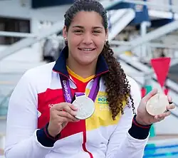 Lorena Miranda trägt eine weiße Trainingsjacke. In der rechten Hand hält sie ihre olympische Silbermedaille, in der linken Hand hält sie eine weitere Medaille mit der Aufschrift London 2012