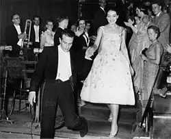 schwarz.weiß Foto aus dem Jahre 1959. Zeigt Maria Callas, wie sie lächelnd im einen Rüschenkleid die Treppe des Concertgebouw vom Frack tragenden Dirigent Nicola Rescigno elegant mit einer haltenden Hand hinunterbegleitet wird. Links sind Mitglieder des Orchesters zu erkennen umrahmt von der anderen Seite der Treppe von applaudierenden Zuschauern.