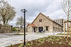 Eine Fotografie, auf der das neue Gelände des Quartiers Sankt Leonhard zu sehen ist. Im Vordergrund steht eine schwarze Laterne. Eine neu angelegte Bepflanzung mit einem grünen Bodendecker und einem jungen Baum mit langsam grüner werdenden Knospen ist im Vordergrund zu sehen. Im Mittelgrund liegt ein mit hell- und dunkelgrauen Platten gepflasterter Platz. Dahinter sind die restaurierten Scheunengebäude zu sehen, die moderne, schwarze Sprossenfenster haben. Eine ebenfalls aus schwarzem Stahl und Glas bestehende Eingangstür ist dem Betrachter zugewandt. Davor stehen vier Menschen an einem Stehtisch mit Husse. Im Hintergrund links sieht man einen Teil der Sankt Leonhard-Straße durch einen steinernen Torbogen hindurch, rechts erhebt sich der moderne und neu gebaute sechsstöckige Flachbau.