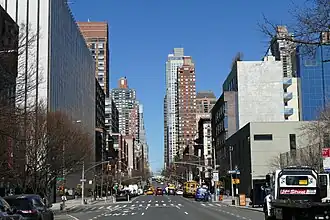 Foto einer mehrspurigen Straße, die von Hochhäusern und modernen Wolkenkratzern gesäumt ist. Auf der Straße herrscht lockerer Verkehr, es sind mehrere gelbe Taxis und ein gelber Schulbus erkennbar