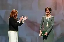 Preisfavoritin rechts im Bild die Bühne betretend und mit Applaus begrüßt von der links stehenden Moderatorin.