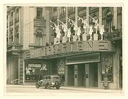 Historisches Foto des Varieté Wintergarten in Berlin, Juni 1929, mit Tänzerinnen auf dem Dach und einem klassischen Auto vor dem Eingang. Das Foto zeigt die Fassade des Wintergarten Varieté-Theaters im Juni 1929. Das Gebäude ist reich verziert, mit kunstvollen architektonischen Details wie gewölbten Fenstern und dekorativen Balkonen. Über dem Eingang prangt der Schriftzug „WINTERGARTEN“ in großen, beleuchteten Buchstaben. Darüber sind Skulpturen von Künstlern zu sehen, die Trompeten spielen, was die künstlerische Atmosphäre des Theaters unterstreicht. Unter dem Schriftzug befindet sich ein weiteres Schild mit der Aufschrift „INTERNATIONALES VARIETÉ“. Der Eingang des Theaters ist sichtbar, flankiert von Plakaten und Werbeanzeigen für die Aufführungen. Vor dem Gebäude steht ein Oldtimer. Die Szene vermittelt die Eleganz und den kulturellen Glanz des Theaters in dieser Ära.