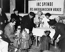 Eine Gruppe von Menschen, darunter Kinder und ältere Erwachsene, versammelt sich um eine Tisch, während eine Frau in einem weißen Mantel Milch in Flaschen ausschenkt; im Hintergrund ein Schild mit der Aufschrift IRC Spende des amerikanischen Volkes.