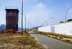 Farbfotografie entlang der Berliner Mauer auf der rechten Seite. Links steht ein Wachturm und in der Mitte ist ein Asphaltweg mit verwilderten Grünflächen. Im Hintergrund sind zwischen den Wolken Fabrikschornsteine und der Fernsehturm zu erkennen.
