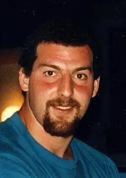 Épalle sitzt seitlich zur Kamera, schaut aber direkt aus dem Bild. Er trägt einen schwarzen Vollbart und einen blauen Pulli.