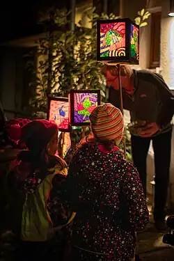 Kinder (von hinten zu sehen) mit bunten Martinslaternen an einer Tür, aus der ein Mann Süßigkeiten reicht