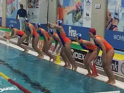 Sieben Niederländerinnen im orangefarbenen Badeanzug springen vom Beckenrand ins Wasser. Die Torhüterin trägt eine rote Bademütze, die Feldspielerinnen tragen eine blaue Kappe.