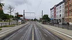 Straße. Mitte: zwei Straßenbahngleise und überdachte Haltestellen auf beiden Seiten. Rechts: drei Autospuren. Links: mindestens eine Autospur.