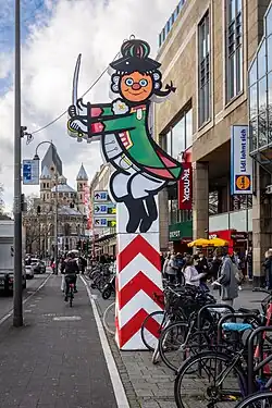 Straßenszene in Köln mit einer großen bunten Karnevalsfigur auf einem rot-weiß gestreiften Sockel am Straßenrand. Die Figur stellt eine stilisierte, lächelnde Person in grün-rotem Uniformkostüm mit Dreispitz und Degen dar – typisch für den Kölner Karneval. Im Hintergrund sieht man Passanten, Fahrradständer, Geschäftsschilder (u. a. Lidl, TK Maxx) und weiter hinten die romanische Kirche Groß St. Martin mit ihren markanten Türmen. Es ist tagsüber, der Himmel ist bewölkt, und es herrscht reger Fuß- und Radverkehr.