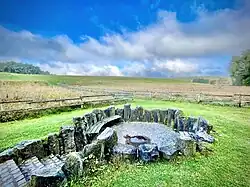 In einer Wiesen bzw. Weidenlandschaft fassen senkrecht gesetzte Basaltsteine eine vertiefte, gepflasterte runde Fläche ein. Darin ein rundes Loch mit rostbraun erscheinendem Wasser