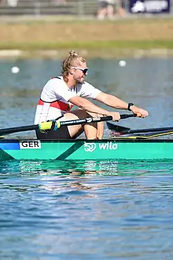 Hagemeister trägt das weiße Nationaltrikot mit dem roten Brustring und eine schwarze Hose. Der Rechtsausleger sitzt im grünen Boot und rudert.
