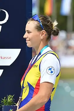 Maria-Magdalena Rusu schaut nach links. Sie trägt den rumänischen rot-gelb-blauen Ruderanzug über einem weißen T-Shirt sowie eine zu den Haaren hochgeschobene Sonnenbrille. Um den Hals hat sie ein blaues Band, an dem die Bronzemedaille befestigt ist. In der linken Hand hält sie einen kleinen Blumentopf.