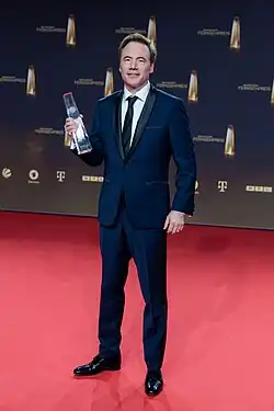 Mann im dunken Anzug vor Sponsorenwand, in der rechten Hand einen Fernsehpreis