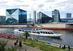 Frontale Farbfotografie auf das Glasgebäude. Direkt an der Uferpromenade stehen zwei sechs geschossige Teilgebäuden mit einer kleineren Glasfront in der Mitte, die als Aufschrift „DB Berlin Hauptbahnhof“ hat. Alle drei Gebäude sind von einer großen Glasröhre durchzogen. An der linken Seite steht ein grauer Turm mit dem DB Logo.