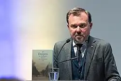 Mittvierziger mit kurzen Haaren und Bart, grauem Anzug, Weste und Krawatte am Rednerpult. Vor ihm aufgestellt ein Buch mit dem Titel „Dnipro. An Entangled History of a European City“. Im Hintergrund blau-gelbe Beleuchtung.