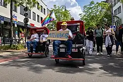 Zwei Rikschas kommen frontal ins Bild, der Passagier der rechten hält ein Pappschild auf dem "miteinander" steht.