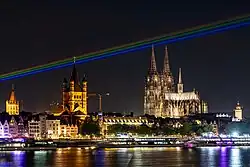 nächtliches Rheinpanorama Kölns (Dom, Groß-St. Martin, Rathaus, Schiffe, Fluß) mit farbigen Strahlen in Regenbogenfarben am Himmel