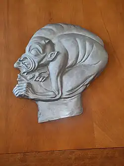 Relief aus glänzendem Silber; Männerkopf im Profil nach links blickend, im Gesicht verbirgt sich eine kauernde Kreatur, die die Augen mit dem Männergesicht gemeinsam hat, der Rücken der Kreatur bildet den Schädel, das untere Gesicht die Nase, der angewinkelte Oberschenkel die Wange, der linke Unterarm den Unterkiefer des Mannes