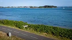 Dolmen aus grob behauenen Granitblöcken direkt am Straßenrand, mit deutlich sichtbarer Deckplatte auf mehreren senkrechten Tragsteinen. Der Steinbau hebt sich aus einem grünen Farnstreifen vor dem dahinterliegenden blauen Meer ab
