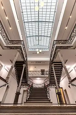 Innenansicht eines historischen Treppenhauses mit einem zentralen Treppenlauf, der in der ersten Etage endet und sich dort in zwei seitliche Treppenarme aufteilt, die in entgegengesetzte Richtungen weiter nach oben führen. Die Treppengeländer sind aus schwarzem, verziertem Eisen mit floralen Mustern und roten Handläufen. Der Raum ist weiß gestrichen und wird durch ein großes Glasdach mit Stahlrahmen hell erleuchtet. Mehrere kugelförmige Lampen hängen von der Decke.