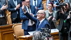 Lächelnder Mann mittleren Alters mit Glatze und Brille in einem dunkelblauen Anzug, der im Parlament inmitten applaudierenden Menschen mit als Geste des Dankes erhobenem Arm steht. Im Hintergrund weitere Personen, die applaudieren, sowie Kameraleute, die das Geschehen filmen.