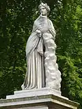 Statue von Polyhymnia im Park Sanssouci, Potsdam, Schrägansicht von vorn links und von unten, da sie auf einem hohen Podest steht; sie stützt sich mit dem linken Unterarm und dem rechten Ellenbogen auf einen halbhohen Baumstumpf, die rechte Hand unter ihrem Kinn; sie trägt einen Umhang