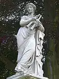 Statue von Euterpe im Park Sanssouci, Potsdam, Schrägansicht von vorn links und von unten, da sie auf einem hohen Podest steht; sie lehnt sich mit ihrer linken Seite gegen einen mit Tüchern verdeckten Gegenstand, in beiden Händen hält sie einen Aulos