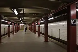 Blick entlang des Mittelbahnsteigs der Bahnhofs 57th Street. Der Raumeindruck wird bestimmt von den cremefarben gefliesten, teils schmutzigen Hintergleiswänden, den massiven dunkel-weinroten T-Trägern, die als Stützenreihen in der Nähe der beiden Bahnsteigkanten verlaufen, von den dunkel angelaufenen Betonplatten des Bahnsteigbodens und von einer dunkelbeige angestrichenen Decke aus quer verlaufenden Stahlträgern und vermutlich Betonfüllungen. Entlang der Decke an den Bahnsteigkanten verlaufen ununterbrochene Bänder von Leuchtstoffröhren, von denen aber manche defekt sind und nicht leuchten. In der Bahnsteigmitte ist eine dritte, allerdings aus Einzelröhren mit jeweils Lücken zwischen den Leuchtstofflampenhaltern bestehende Lichtreihe. Im Bildhintergrund blickt man direkt auf zwei jeweils die halbe Bahnsteigbreite einnehmende Treppen, die vom Bahnsteig nach oben führen. An jeder zweiten der dunkelroten Bahnsteigstützen sind Schilder mit dem Bahnhofsnamen montiert im typischen Transit-Authority-Design in der Schreibweise „57 Street“ mit weißer Schrift auf schwarzem Grund. An den Hintergleiswänden ist der Bahnhofsname in regelmäßigen Abständen aufgedruckt in der Schreibweise „57th Street“.