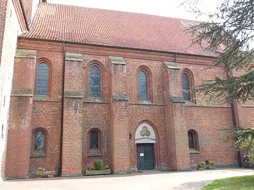 Nördliches Seitenschiff, Fas­sade zwei­geschos­sig und Fenster­formen wie Südseite, gotisches Portal mit ersetz­tem char­riertem Tympanon