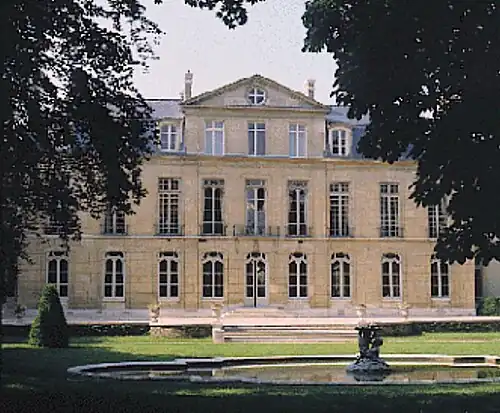 Blick aus dem Garten über ein kleines Wasserbassin auf die dreigeschossige Fassade des Hôtels de Castries mit vielen hohen Fenstern und einem mittig positionierten Dreiecksgiebel auf Dachgeschosshöhe. Über die gesamte Breite ist dem Gebäude eine Terrasse vorgelagert, zu der eine breite Treppe mit nur wenigen Stufen hinaufführt. Auf der Terrassenbrüstung stehen weiße Steinvasen. Am rechten und linken Bildrand sind dicht belaubte Bäume zu sehen.