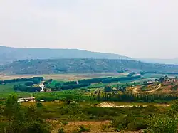 Blick von einer Anhöhe über recht karge Landschaft mit einigen Feldern und Häusern auf einen Fluss. Dieser hat einen sich schlängelnden, ruhigen Verlauf und braunes Wasser. Entlang der Uferkante stehen in einer ordentlichen, aber teils unterbrochenen Linie niedrige, recht kugelförmige Bäume dicht beisammen. In der Ferne hinter dem Fluss sind Hügel.