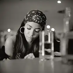 Aaliyah schwarz-weiße Fotografie, die an einem Tisch oder an Klavier sitzt. Sie trägt ein Bandana mit floralem Muster und große Creolen-Ohrringe. Das lange Haar der Person ist sichtbar, und sie scheint ein ärmelloses Oberteil zu tragen. Auf dem Tisch vor der Person befinden sich mehrere Gläser und Flaschen, die ebenfalls etwas unscharf sind. Im Hintergrund ist eine verschwommene Umgebung mit einigen Lichtquellen sichtbar, die auf einen schwach beleuchteten Raum hinweist, möglicherweise ein Restaurant oder eine Bar.