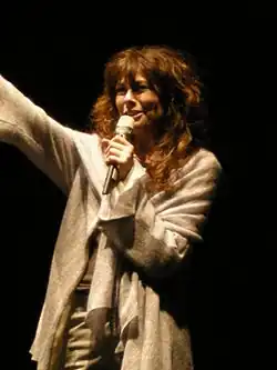 Alice mit langem, lockigen, braunen Haar steht auf der Bühne und spricht ins Mikrofon. Sie trägt einen grauen, offenen Cardigan und hat einen freundlichen Gesichtsausdruck. Der Hintergrund ist dunkel. In der einen Hand hält Alice ein Mikrofon, während sie mit der anderen Hand eine Geste macht, als würde sie jemanden zuwinken. Das Haar fällt natürlich über die Schultern.