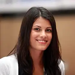 Alisar Ailabouni (2010) mit langen, dunklen Haaren und lächelndem Gesicht, trägt einen weißen Blazer. Der Hintergrund ist unscharf und lässt keine spezifischen Details über die Umgebung erkennen.