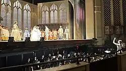 Im Hintergrund eine Opernbühne mit Mitwirkenden, im Vordergrund ein Orchester im Orchestergraben, dirigiert von Alma Deutscher.