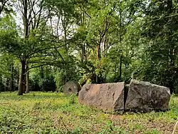 Ein etwa 60 cm hoher, in vier Teile gespaltener und lose zusammengelegter Steinblock in einem lichten Wald, dahinter ein Mühlstein, der hochkant steht.