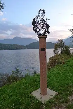vor einem See steht eine hohe Stele, auf dieser ist eine Büste von Preuß, im Hintergrund Berge