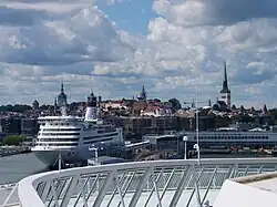 Farbfotografie über eine moderne Brücke zum Kreuzfahrtschiff an der Anlegestelle. Im Hintergrund Häuser und Kirchtürme.