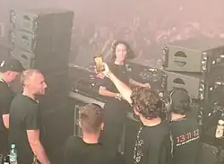 Amelie Lens steht im Mittelpunkt des Bildes an verschiedenen DJ-Playern, hinter ihr eine Menschenmenge und im Vordergrund stehen einzelne Menschen. Sie hat langes schwarzes Haar und trägt schwarze Kleidung.