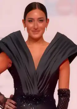 Amina Khalil mit langen, braunen Haaren, die an den Enden heller werden. Sie trägt eine Halskette mit mehreren Ketten und kleine, runde Ohrringe. Im Hintergrund ist eine unscharfe Außenszene erkennbar, die Grünflächen und möglicherweise eine Gebäudestruktur durch ein Fenster zeigt. Sie hält ein pinkfarbenes Smartphone am Ohr. Das Bild vermittelt den Eindruck eines nachdenklichen oder interaktiven Moments, da Amina Khalil scheinbar auf das Telefongespräch fokosiert ist.