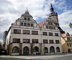 Altes Gebäude, weiß verputzt, zahlreiche dunkle Steinbalken gliedern die Fassade, zwei Spitze Giebel bilden die Front zum Marktplatz.