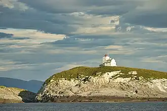 Foto einer grasbedeckten Insel mit einem weißen Leuchtturm darauf und bewölktem Himmel im Hintergrund