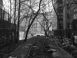 Schwarzweißaufnahme eines alten Bahnsteigs in einem Einschnitt. Beengt zwischen mehrstöckigen Wohnhäusern rechts und links wirken die aus Beton gefertigten, teils mit Schutt und Unrat aufgefüllten ehemaligen Gleisbetten und der Bahnsteig wie vergessen. Im Bildhintergrund fällt der Blick nahtlos auf einen kleinen Teil der Fassade eines weiteren großen Gebäudes, des Yankee Stadiums.