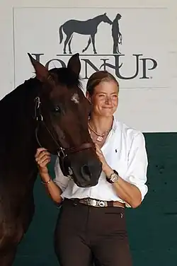 Andrea Kutsch trägt eine braune Hose und ein weißes Oberteil. Sie hat blondes zusammengebundenes Haar und streichelt ein braunes Pferd am Kopf.