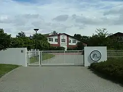 Im Hintergrund ist ein Haus zu sehen, im Vordergrund befindet sich ein Zaun mit verschlossenem Tor zur Einfahrt.