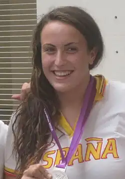 Andrea Blas trägt ein weißes Polohemd mit der gelben Aufschrift Espana. Trainingsjacke. Mit der rechten Hand hält sie ihre olympische Silbermedaille, die an dem lilafarbenen Band um den Hals von Blas befestigt ist.