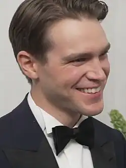 Andrew Burnap (2021) bei den 74th Tony Awards trägt einem eleganten, dunklen Smoking mit einer weißen Anzugbluse und einer schwarzen Fliege. Die Haare sind ordentlich zur Seite gekämmt. Im Hintergrund ist eine neutrale, helle Wand zu sehen, die schlicht gehalten ist. Auf der rechten Seite des Bildes ist eine kleine grüne Pflanze sichtbar, die etwas Lebendigkeit in die Szene bringt.